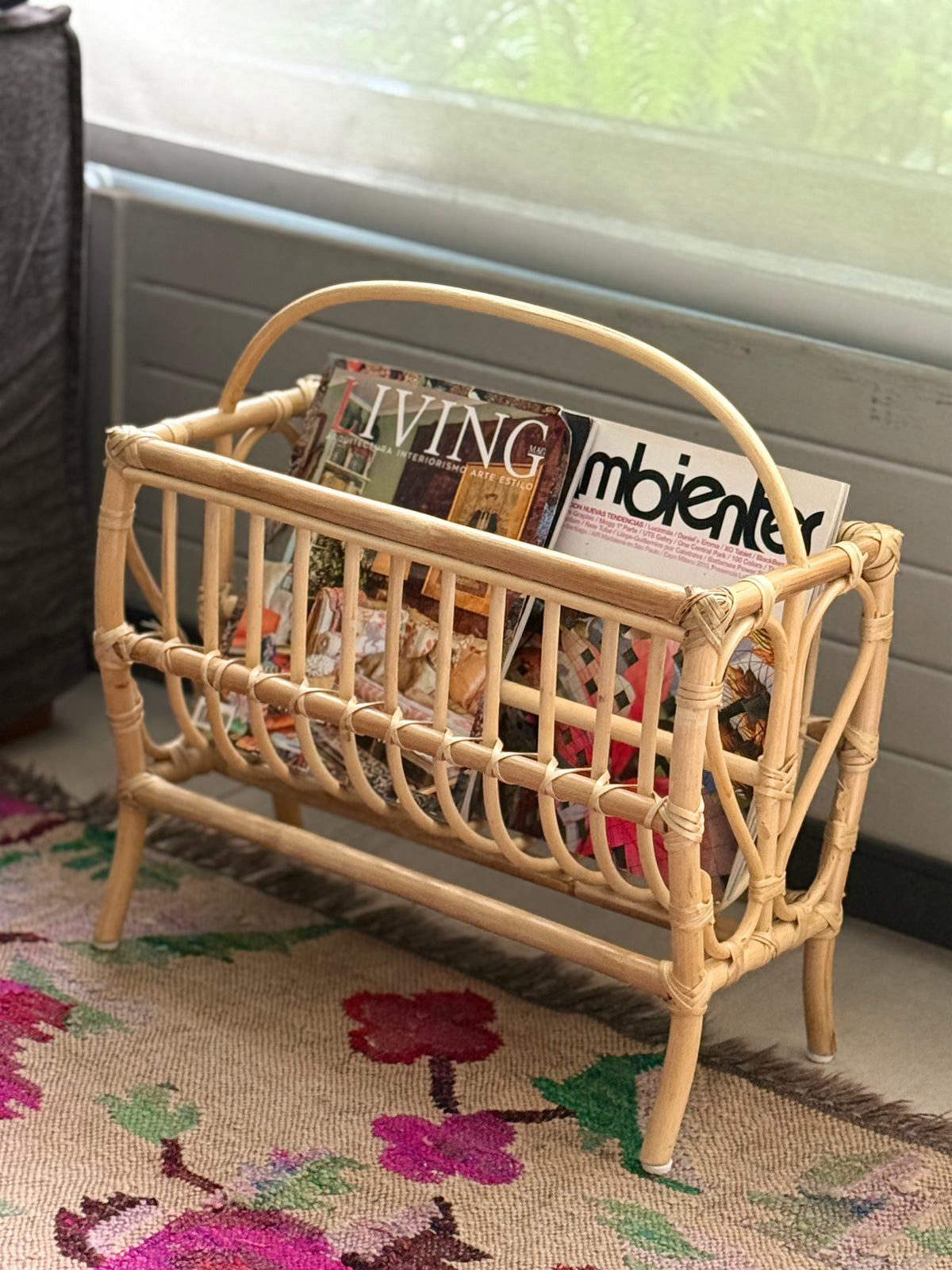 Revistero de bambú con revistas, sobre una alfombra junto a una ventana